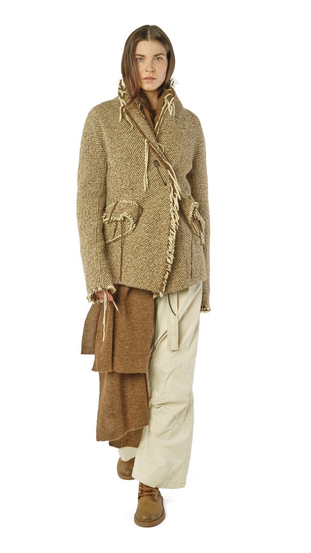Frau trägt oversized beige Strickjacke mit Fransen, weite beige Hose und braune Stiefel, hält braune Tasche.