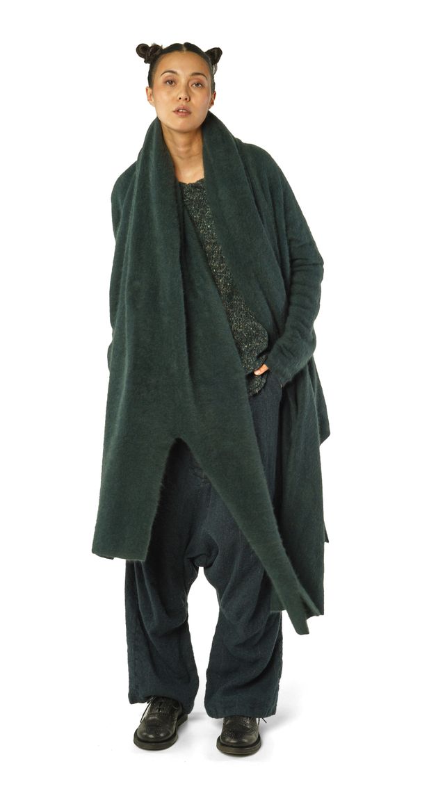 Person trägt oversized grünen Wollmantel mit weitem Schnitt, schwarze Hose und Sneaker, lässige Pose.